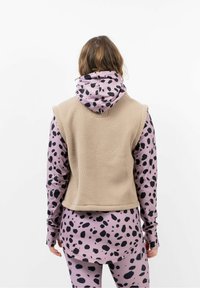 Hellbraunes Fleece-Weste über einem rosa Langarmshirt mit schwarzen Punkten, das lange Ärmel und einen abgerundeten Saum hat, von hinten gesehen.
