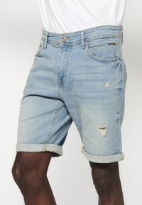 Shorts en denim bleu clair avec une coupe décontractée, présentant des détails usés, des poignets retroussés et un design à cinq poches. Fermeture à bouton métallique.