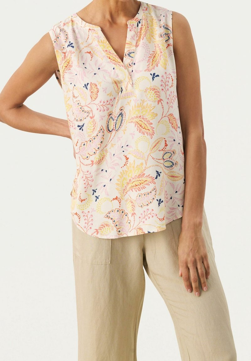 Blouse sans manches rose clair avec un col en V, présentant un motif floral jaune, orange et bleu. Portée avec un pantalon en lin beige.