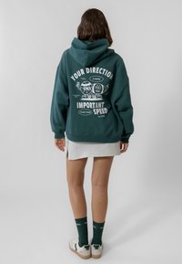 A zöld kapucnis pulóver pamut keverékből készült, fehér grafikus szöveggel és mese figurákkal a hátán. Szürke szoknyával, zöld zoknikkal és sportcipővel párosítva.