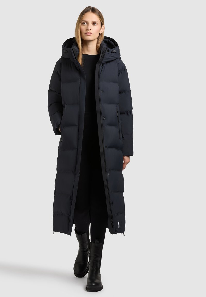 Langer, gepolsterter Mantel aus dunkelblauem Stoff mit Kapuze, Druckknopfverschluss und Seitentaschen. Kombiniert mit schwarzen Stiefeln und einem schwarzen Outfit.