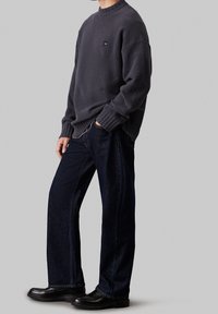 Szary sweter z polaru z prążkowanymi mankietami, połączony z ciemnoniebieskimi, szerokimi spodniami jeansowymi i czarnymi butami. Na swetrze widoczna jest charakterystyczna czarna naszywka z logo.