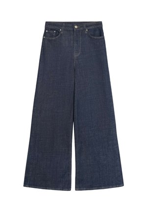 Femme blonde debout, bras croisés, portant un sweat court bleu et un jean large foncé sur fond rouge.
