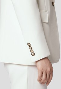 Rękaw białej marynarki z gładką teksturą, czterema złotymi guzikami oraz dopasowanym krojem z dyskretną kieszonką.