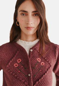 Maroon neulecardigan, jossa on vaaleanpunaisia kukkaompeluja, teksturoitu pinta, pyöreät napit ja V-malli.