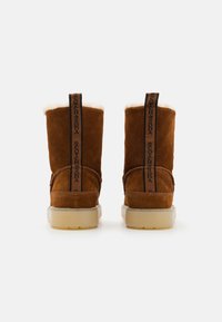 Stivali alla caviglia in suede marrone con fodera in finta pelliccia bianca, suola trasparente e un etichetta con logo nera sul retro. Design semplice e funzionale.
