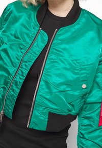 Groene satijnen bomberjack met een zwarte ribgebreide kraag en manchetten, zilveren rits en een linkerzak met knoopdetail. De binnenkant heeft een gequilt patroon.