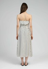Robe midi sans bretelles en tissu blanc avec des pois noirs. Taille élastique, jupe fluide, texture lisse. Portée avec des sandales noires à enfiler.