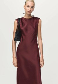 Kvinna iklädd ärml dress i burgundy satin och långa sfäriska örhängen, bärande på en svart vävd axelväska, stående mot en enkel bakgrund.