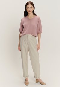 Giovane donna in piedi, che indossa un maglione leggero rosa chiaro a V, pantaloni beige con risvolto e scarpe beige con tacco basso su uno sfondo neutro.
