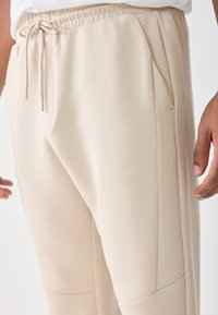 Nærbilde av person som har på seg beige joggebukser med elastisk midje og snor, som viser høyre hånd og side-lomme.