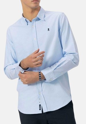 Camisa de manga larga azul claro, con cuello abotonado, y un pequeño logo de pulpo negro en el pecho. Material de algodón, corte relajado, con puños abotonados.