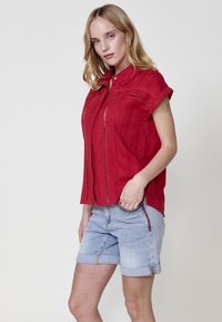 Rode kortemouwige overhemd met knopen en uitsnijdingen; gecombineerd met lichtblauwe opgerolde spijkerbroekshorts met decoratieve stiksels en zakken.