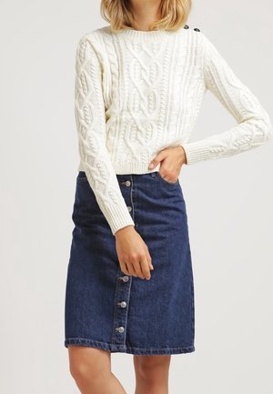 Kvinde iført en hvid kabelstrikket sweater med skulderknapper og et mørkeblå denimskørt med knapper foran.