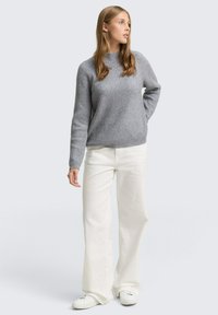 Grauer gerippter Pullover mit Rundhalsausschnitt, kombiniert mit weißen weit geschnittenen Hosen und weißen Sneakers, vor einem neutralen Hintergrund.