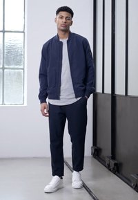 Casaco de bombardeiro azul-marinho sobre uma camisa cinza, combinado com calças azul-marinho e ténis brancos. Tecido suave, design minimalista e corte ajustado.