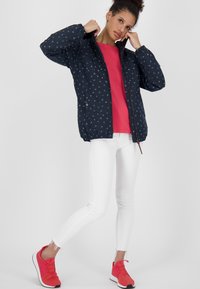 Giovane donna che aggiusta il colletto di un giacca blu navy con piccoli pois bianchi, indossando una camicia rossa, jeans bianchi attillati e sneakers rosse, in piedi su una gamba.