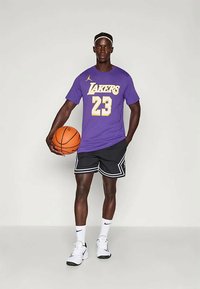 Vijolična majica s napisom "LAKERS" in številko "23" v rumeni barvi, črne kratke hlače z belimi detajli, drži košarkaško žogo, obut v bele superge.