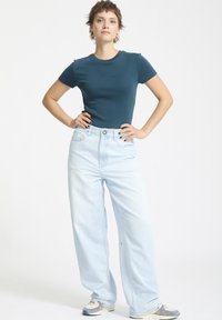 Türkises, tailliertes T-Shirt, hochgeschnittene, weit geschnittene, hellblaue Jeans und graue Sneaker. Verwirrte Haare, Arme in die Hüften gestemmt, neutraler Hintergrund.