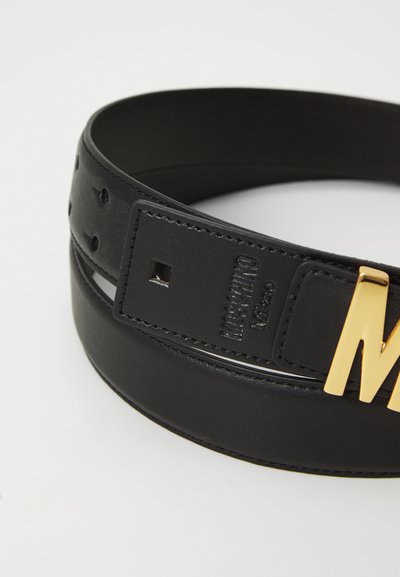 Cinturón de cuero negro con acabado brillante, que presenta una gran hebilla en tono dorado con la letra "M", así como costuras visibles y agujeros para ajuste.