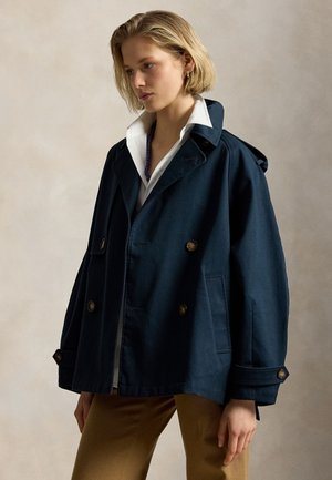 Jeune femme aux cheveux blonds courts portant une veste croisée bleu marine, une chemise blanche à col et un pantalon marron, debout devant un fond neutre.