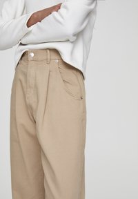 Pantalones beige de cintura alta con pierna recta, confeccionados en tela texturizada. Presentan pliegues sutiles y un único botón de metal a la cintura.