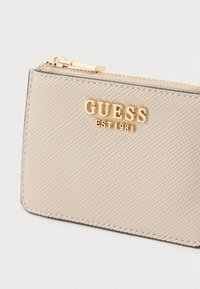 Portofel sintetic bej texturat, cu logo auriu embossat „GUESS EST 1981”, fermoar și cusături contrastante pe margini.