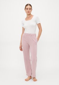 Vrouw die blootsvoets staat, gekleed in een aansluitend wit T-shirt met korte mouwen en losse roze elastische broek met een tailleband, met haar handen in haar zakken tegen een witte achtergrond.