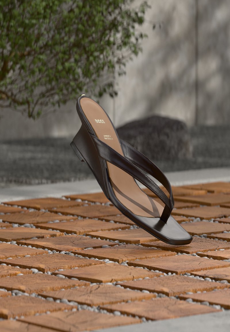 Μαύρο σφήνωμα σανδάλι με λουριά σφήνας τοποθετημένο σε ξύλινη σανίδα και πέτρινο έδαφος σε εξωτερικό χώρο κοντά σε πράσινα φυτά και τοίχο από σκυρόδεμα.