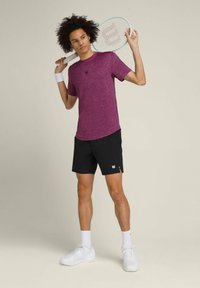 Männliches Sportoutfit mit einem strukturierten, lila kurzärmeligen Shirt und schwarzen Shorts mit einem weißen Logo, kombiniert mit weißen Sneakers und Handgelenkbändern.
