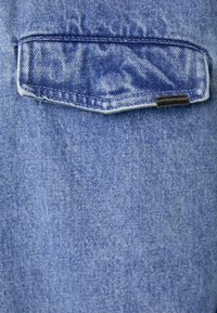 Camisa de ganga com uma cor azul desbotada, apresentando um bolso cosido nas costas e uma pequena etiqueta da marca; textura suave com um design clássico.