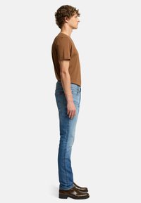 T-shirt in cotone marrone, jeans skinny blu chiaro con texture sbiadita e scarpe in pelle lucida marrone. Vista laterale che mostra la vestibilità e le proporzioni.