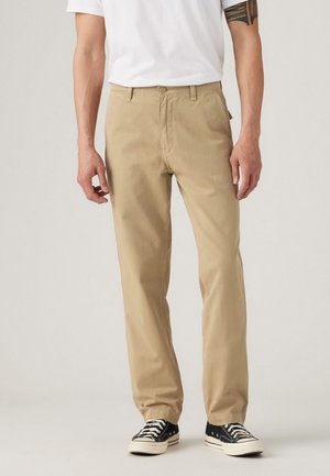 Beige pants made of cotton twill, straight-leg design with a front button, and side pockets. Worn with black sneakers featuring white laces.