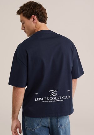 Homme portant un T-shirt oversize bleu marine avec le texte blanc « The Leisure Court Club » dans le dos et un jean bleu clair, de dos.
