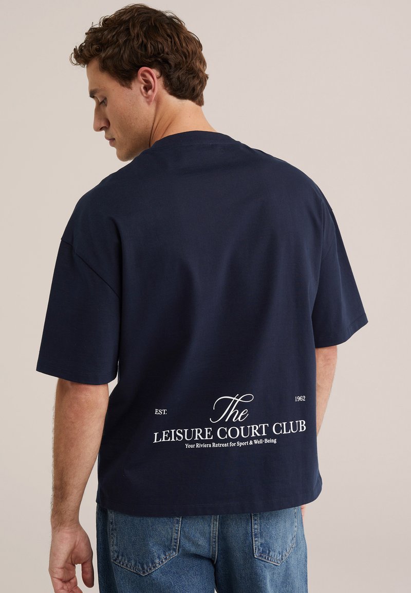 Férfi sötétkék, bő fazonú pólóban, amely hátul fehér felirattal "The Leisure Court Club", és világoskék farmerral, háttal a nézőnek.