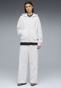 Νεαρό άτομο που φοράει ανοιχτό γκρι φαρδιά ζακέτα με φερμουάρ και ταιριαστό φαρδύ παντελόνι φόρμας, στέκεται μπροστά σε απλό γκρι φόντο.