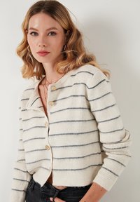 Tej cardigan szürke vízszintes csíkokkal, kerek gallérral és három gombbal. Puha, textúrált anyag, enyhén oversized ujjakkal.