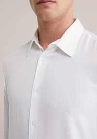 Camicia bianca con colletto classico, caratterizzata da una consistenza liscia e due bottoni visibili sulla patta. Vista ravvicinata del tessuto.