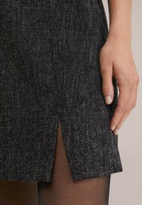 Jupon noir texturé mettant en avant un motif tissé subtil, avec une fente latérale et une coupe ajustée, associé à des collants noirs transparents.