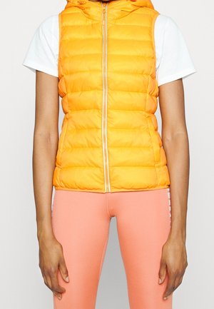 Person wearing a sleeveless yellow puffer vest over a white t-shirt and coral pink leggings, standing against a plain light background.