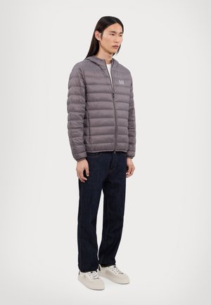 Giacca a vento grigia con cuciture orizzontali, chiusura con zip e polsini elasticizzati, abbinata a jeans di jeans scuri e sneaker chiari.