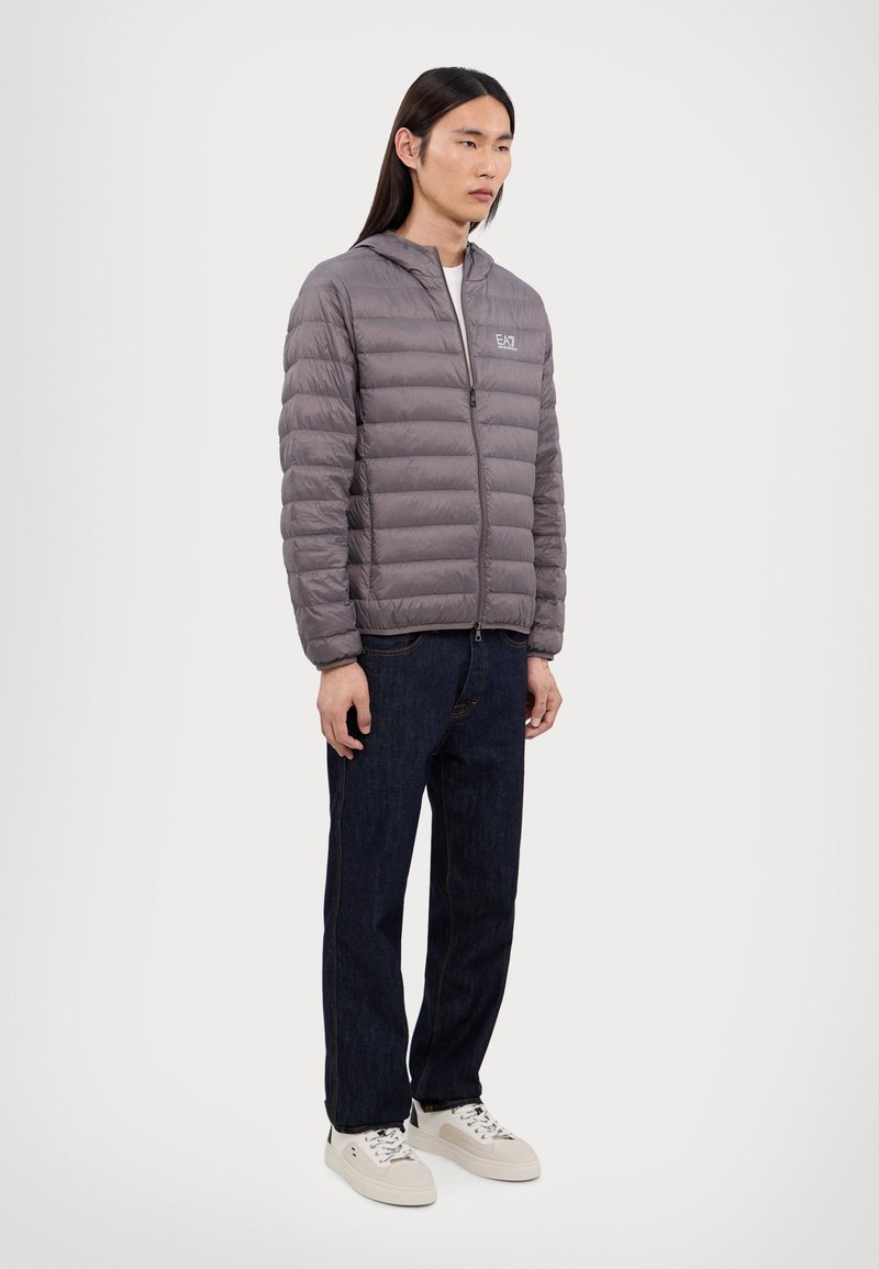 Grå pufferjakke med horisontale sømmer, glidelås, og elastiske mansjetter, kombinert med mørke jeans og lyse sneakers.