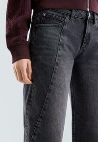 Jeans in denim nero con silhouette tapered, dotati di una struttura a cinque tasche e leggeri effetti di scoloritura. Un manica texturizzata di colore bordeaux è visibile sopra.