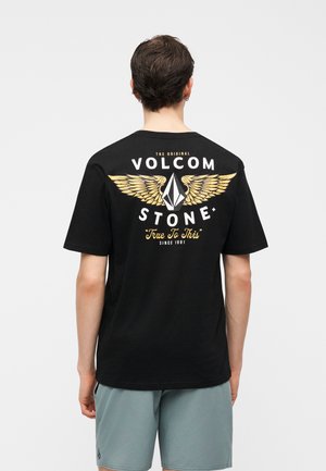 Νεαρός άνδρας, φωτισμένος από πίσω, φοράει μαύρο T-shirt Volcom Stone με χρυσά φτερά και γκρι σορτς σε μονόχρωμο φόντο.