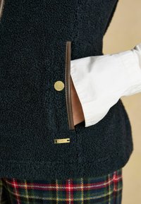 Marineblaues Fleece-Weste mit strukturiertem Oberflächen, braunen Akzenten, goldenen Hardware-Details, seitlichen Taschen und einem darunter sichtbaren weißen Hemd.
