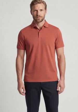 Man staat met een roestkleurig poloshirt met korte mouwen en donkere broek tegen een effen achtergrond.