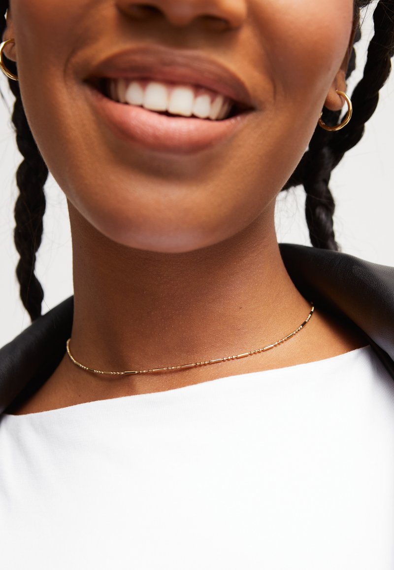 Collier ras du cou en or avec des perles fines et allongées ainsi que de petits accents ronds, porté sur un cou contre une veste noire et un haut blanc.