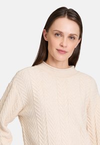Beige kötött pulóver halszirmos mintával, magas nyakkivágással és enyhén laza ujjal, puha, texturált anyagból készült.