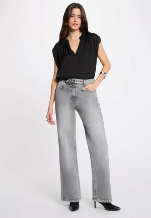 Donna che indossa una camicetta nera senza maniche, jeans grigi a gamba larga a vita alta, tacchi neri con punta e braccialetto d'argento, in piedi con una mano in tasca.