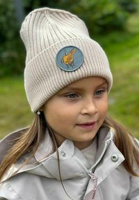 Vaaleanbeige neulepipo, jossa on ribbikuviointi ja pyöreä sininen merkki, jossa on kuvitettu lehmä. Pipoa käyttää lapsi, jolla on pitkät ruskeat hiukset.